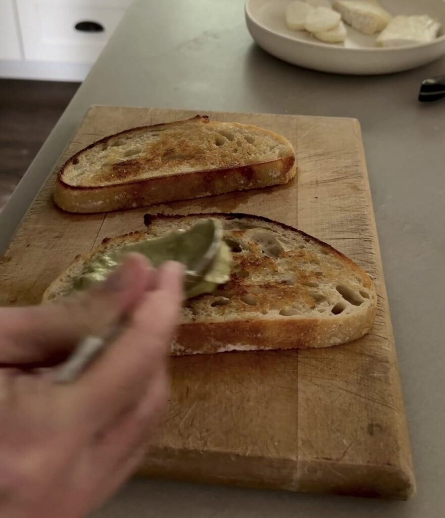 Spreading basil pesto on toasted sourdough for a turkey brie melt
